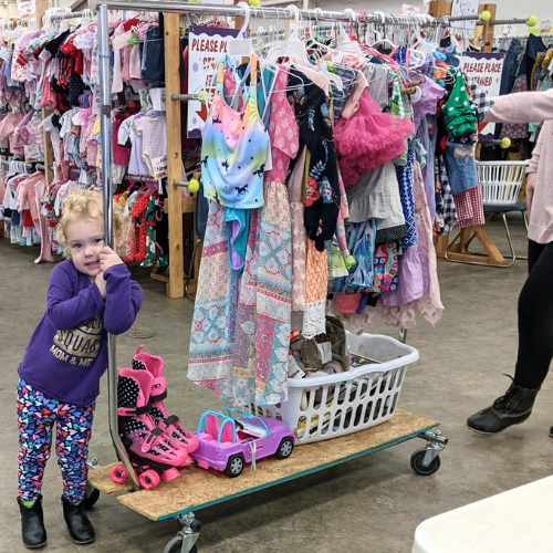 A mom with a large JBF shopping bag on her shoulder stands beside her husband who wears their toddler at a JBF sale.